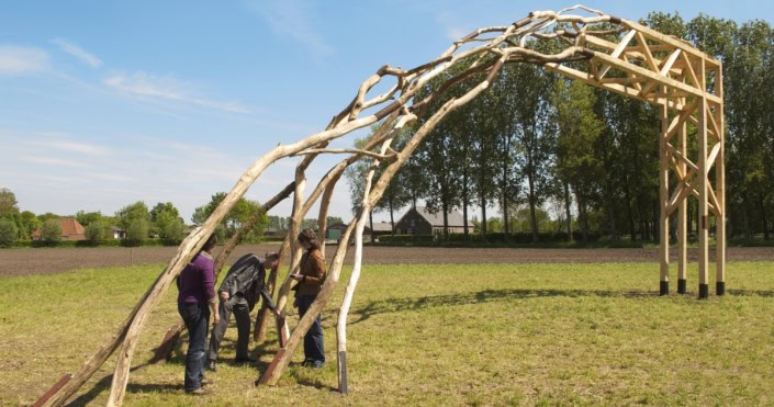 Alt tekst: Mensen bouwen een boombuiltunnel van natuurlijke takken op een grasveld.